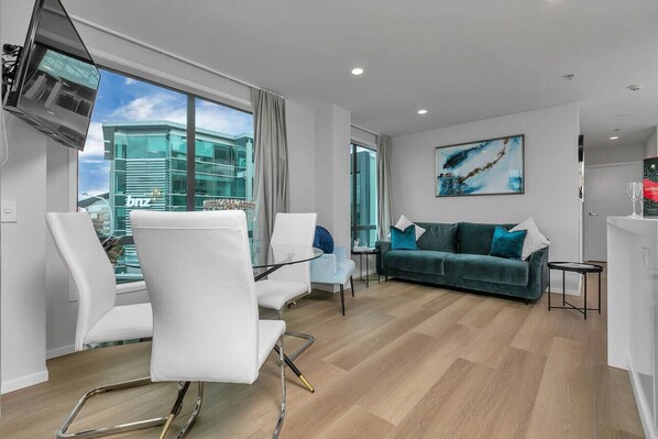 Un salon moderne avec une table en verre, des chaises blanches, un canapé de couleur turquoise et une grande fenêtre donnant sur un immeuble.