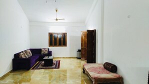 Un salon avec un canapé violet, une petite table, un tapis à motifs, une porte en bois et une fenêtre ornée d’un cadre décoratif.
