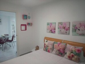 Quarto com cama, uma cadeira e uma mesa. Há decorações nas paredes e uma porta que dá para outro cômodo.