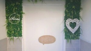 Une porte blanche, ornée d’une pancarte sur laquelle on peut lire « Bonjour » et, à sa gauche, d’une autre pancarte avec le mot « Bienvenue », le tout entouré de feuillage verdoyant.