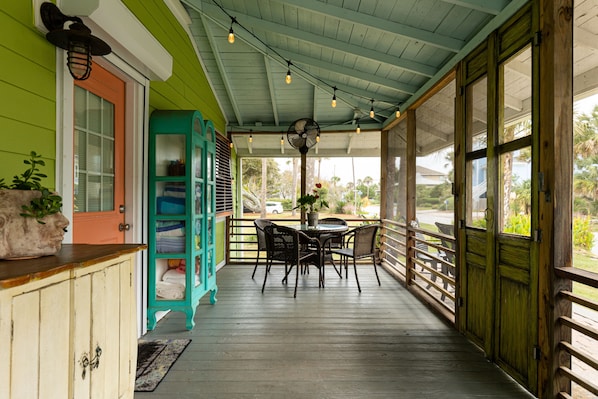 A covered porch with a wooden table and chairs, a teal bookshelf, and a large window with a view of the outdoors.