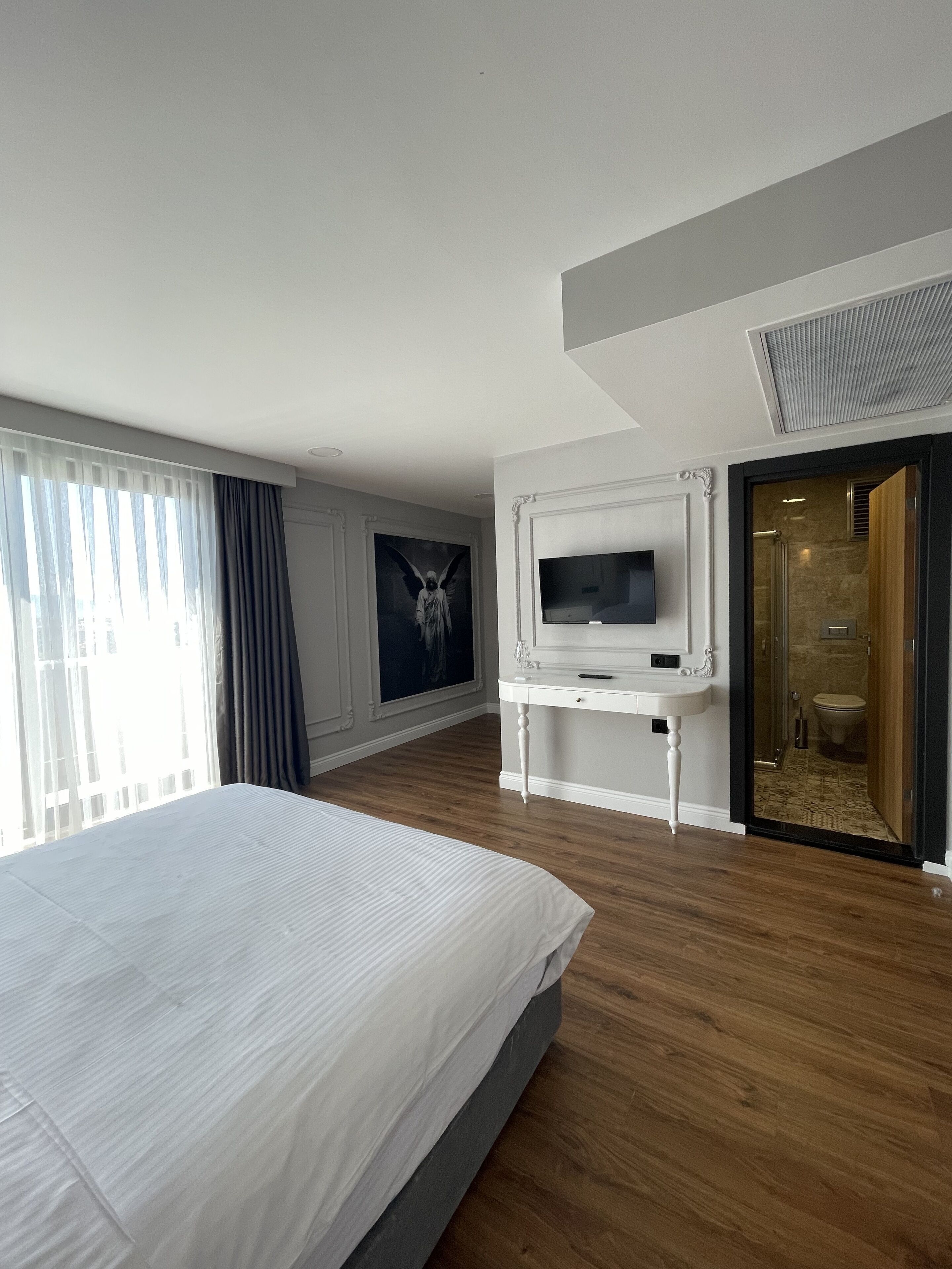 Une chambre d’hôtel avec un grand lit, un téléviseur posé sur un meuble blanc, un tableau accroché au mur et une salle de bain visible à travers une porte ouverte.