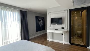 Habitación de hotel con una cama grande, un televisor sobre un soporte blanco, un cuadro en la pared y un baño visible a través de una puerta abierta.