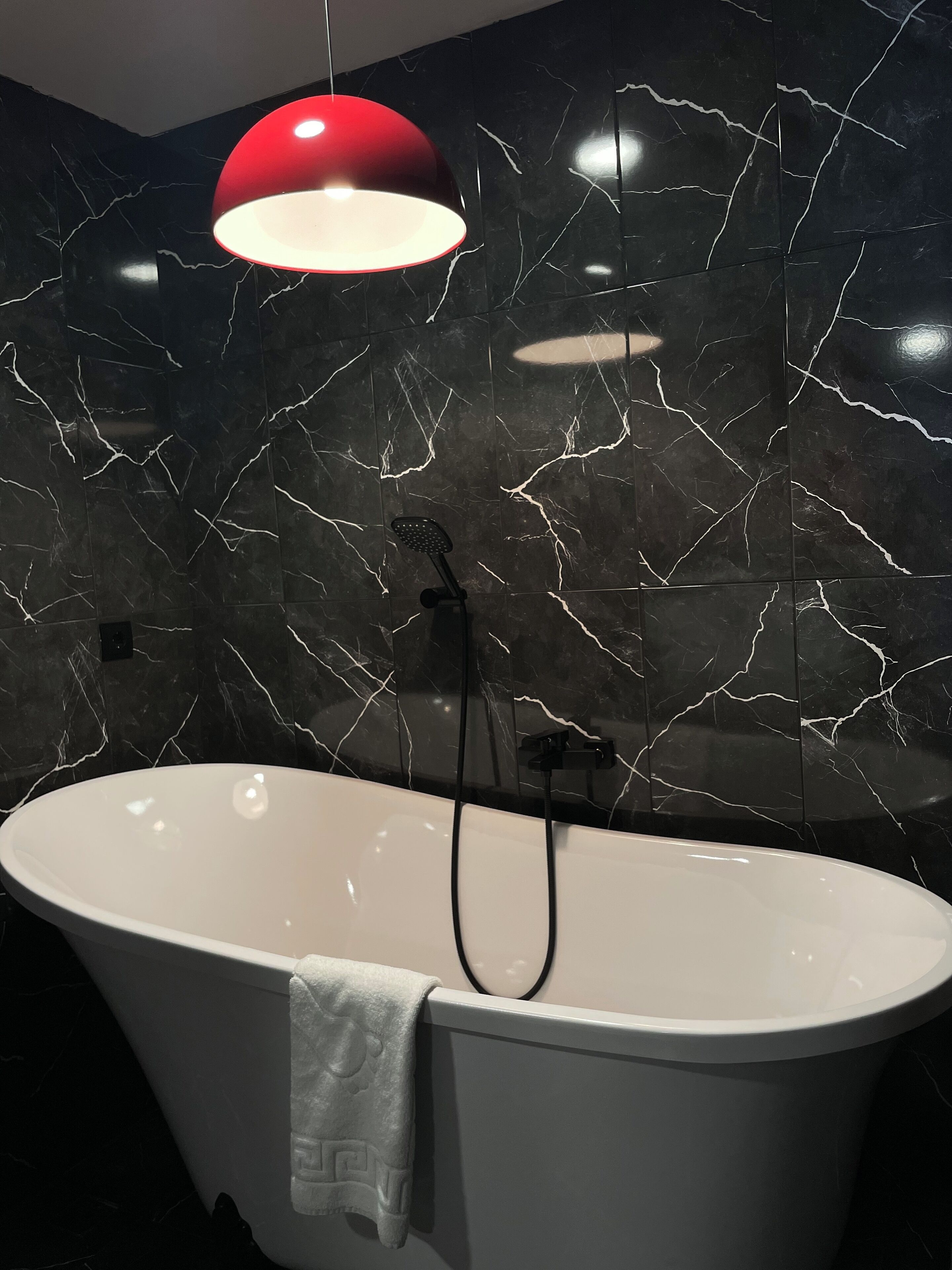 A modern bathroom with a freestanding bathtub, a red pendant light, and black marble walls.