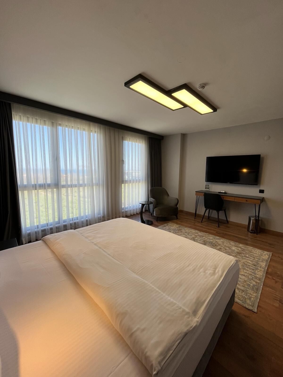 Una habitación de hotel moderna con una cama grande, un escritorio con silla, televisión y vistas al exterior a través de amplios ventanales.