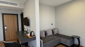 Ein moderner, kompakter Wohnraum mit Holzboden, einem grauen Sofa, einem kleinen Tisch, einem an der Wand befestigten Fernseher und einer Deckenleuchte.