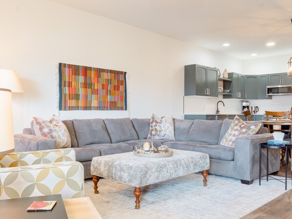 Uma sala de estar moderna com um sofá modular cinza, uma poltrona estampada e uma mesa de centro. Um quadro abstrato e colorido está pendurado acima do sofá.