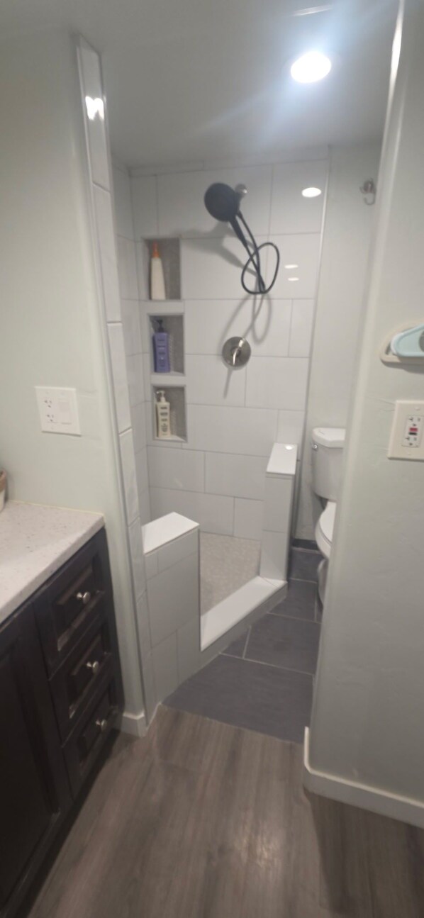 A narrow bathroom with a shower, toilet, and a small shelf with toiletries. - UofA Tiny House + Fire Pit (Tucson)