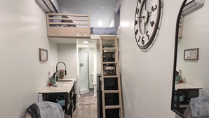 A compact room with a bunk bed, a small kitchenette, a ladder, and a large wall clock. - UofA Tiny House + Fire Pit (Tucson)