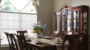 A dining room with a wooden table set for six, surrounded by six chairs. A chandelier hangs from the ceiling, and a large window with blinds is visible.