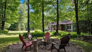 Une cabane dotée d’un porche abrité, entourée d’une forêt. On aperçoit, au premier plan, des fauteuils Adirondack et un foyer.