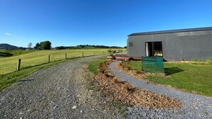 A gravel path leading to a modern building with a metal exterior, surrounded by a fenced field and open landscape.