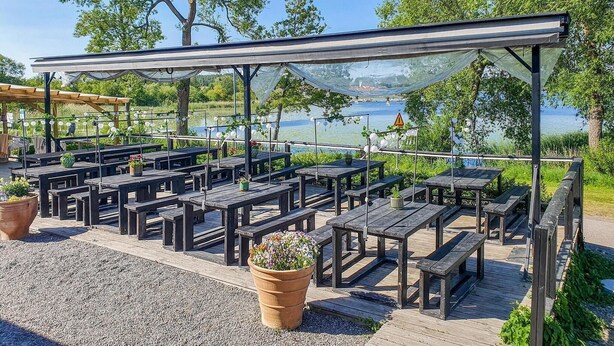 An outdoor seating area with wooden tables and benches, a pergola with a glass canopy, and a view of a lake. - Beautiful home in Gnesta with WiFi (Gnesta)