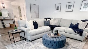 A modern living room with a grey sectional sofa, a round ottoman, a coffee table, and a grey rug. There are framed pictures on the wall and a lamp on a side table. - Modern Home with Private Pool (Phoenix)