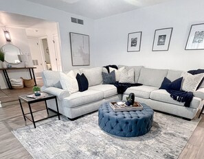 Ein modernes Wohnzimmer mit einem grauen Ecksofa, einem runden Hocker, einem Couchtisch und einem grauen Teppich. An den Wänden hängen Bilderrahmen und auf einem Beistelltisch steht eine Lampe.