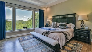 Une chambre à coucher avec un grand lit, un banc, une table de chevet, une lampe et une vue sur l’extérieur.