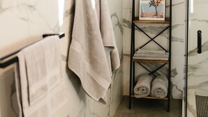 A bathroom with marble walls, a glass shower enclosure, a towel rack, and a shelf with towels and a framed picture. - Joshua Tree Monochrome - 5 mins from National Park (Joshua Tree)