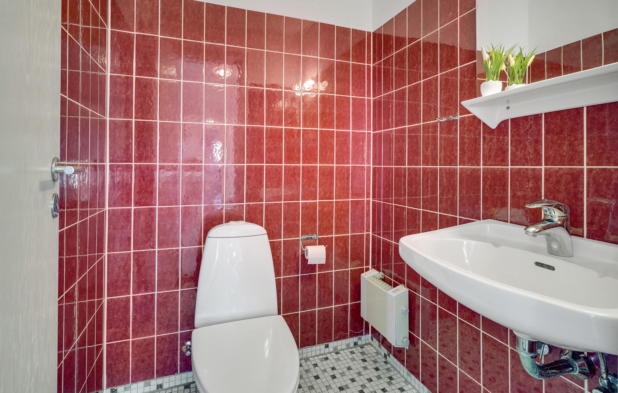 A bathroom with red tiles, a white toilet, a white sink, and a small shelf with plants.