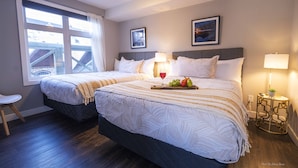 A hotel room with two beds, a window with curtains, a small table with a lamp, and a framed picture on the wall.