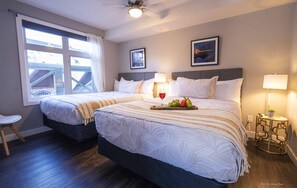 Quarto de hotel com duas camas, uma janela com cortinas, uma mesinha com abajur e um quadro pendurado na parede.