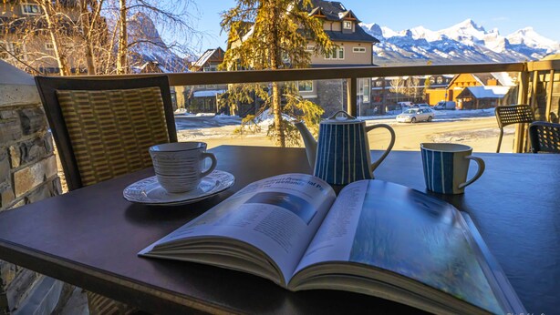 A table with an open book, two cups, and a teapot on a balcony overlooking a snowy mountain landscape.
