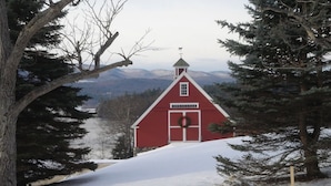 Ein rotes Bauernhaus mit Wetterfahne, umgeben von schneebedecktem Boden und immergrünen Bäumen.