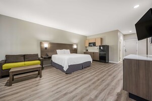 Una habitación de hotel moderna con cama, sofá, banca, kitchenette y televisor de pantalla plana.