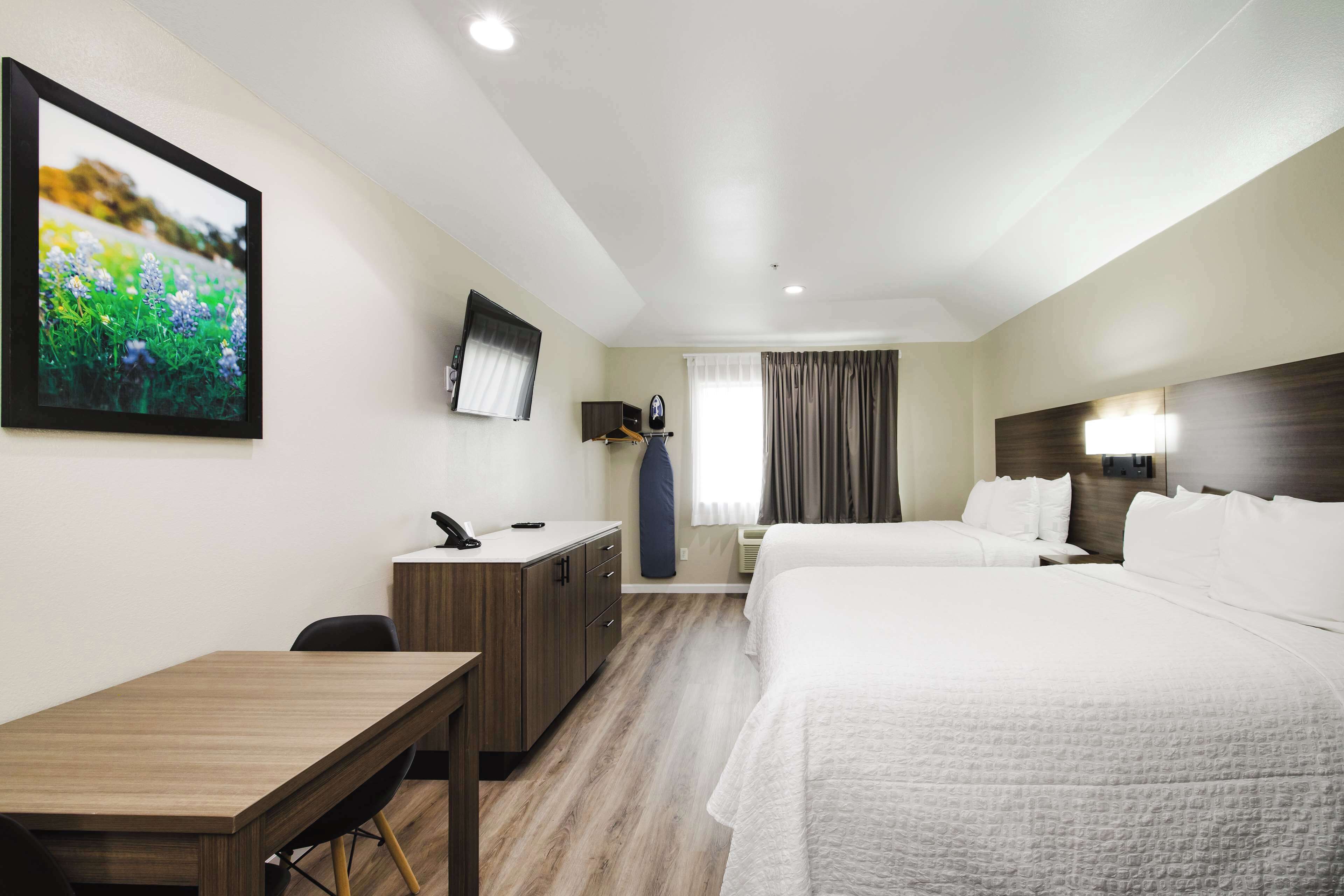 A hotel room with two beds, a desk, a TV mounted on the wall, and a window with curtains.