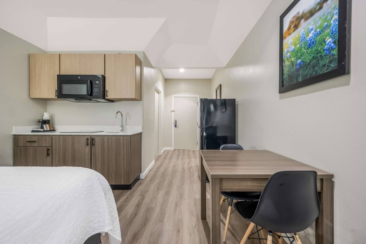 Een kleine kamer met kitchenette, een bed, een eettafel met stoel, en een lijst met een afbeelding aan de muur.