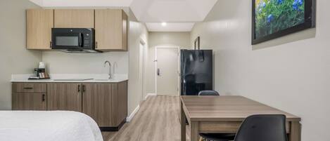A compact room with a kitchenette, a bed, a dining table with a chair, and a framed picture on the wall.