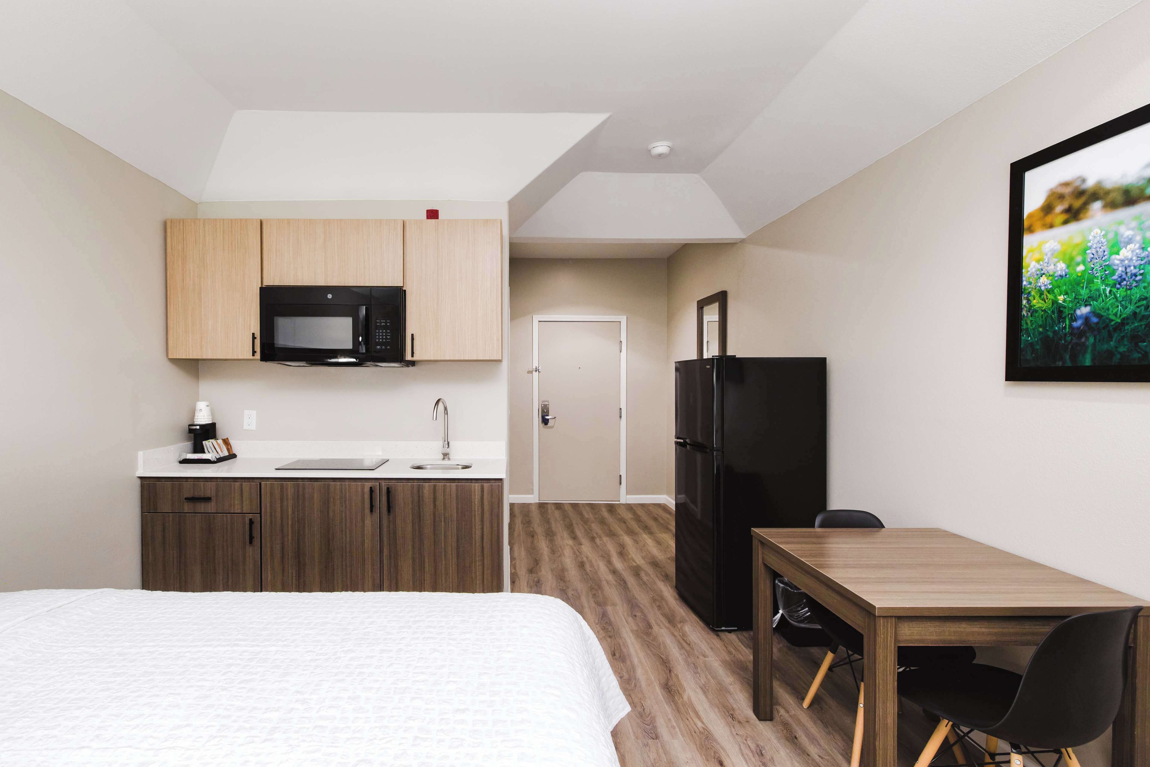 A hotel room with a bed, a kitchenette with a microwave, a refrigerator, a dining table with chairs, and a door leading to another room.