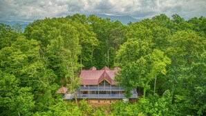 A two-story lodge with a red roof nestled among dense forest, surrounded by mountains.