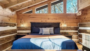 A log cabin bedroom with a bed, two bedside lamps, and a view through the windows.