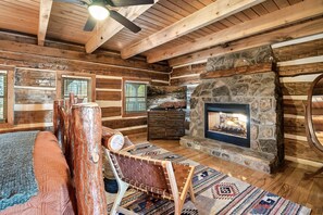 A log cabin interior with a stone fireplace, wooden beams, a large window, and a rustic wooden bench.