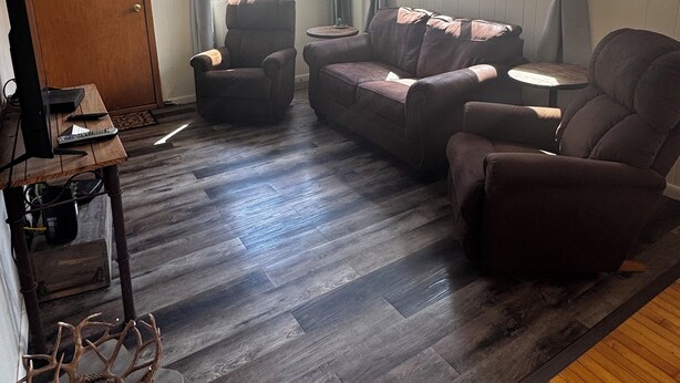 Un salon avec un parquet en bois foncé, un canapé marron, un fauteuil marron, une petite table ronde et une lampe.