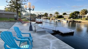 A waterfront patio with blue Adirondack chairs, a dock, and a calm water body. - Beautifully remodeled in 2025. Gulf access. (New Port Richey)
