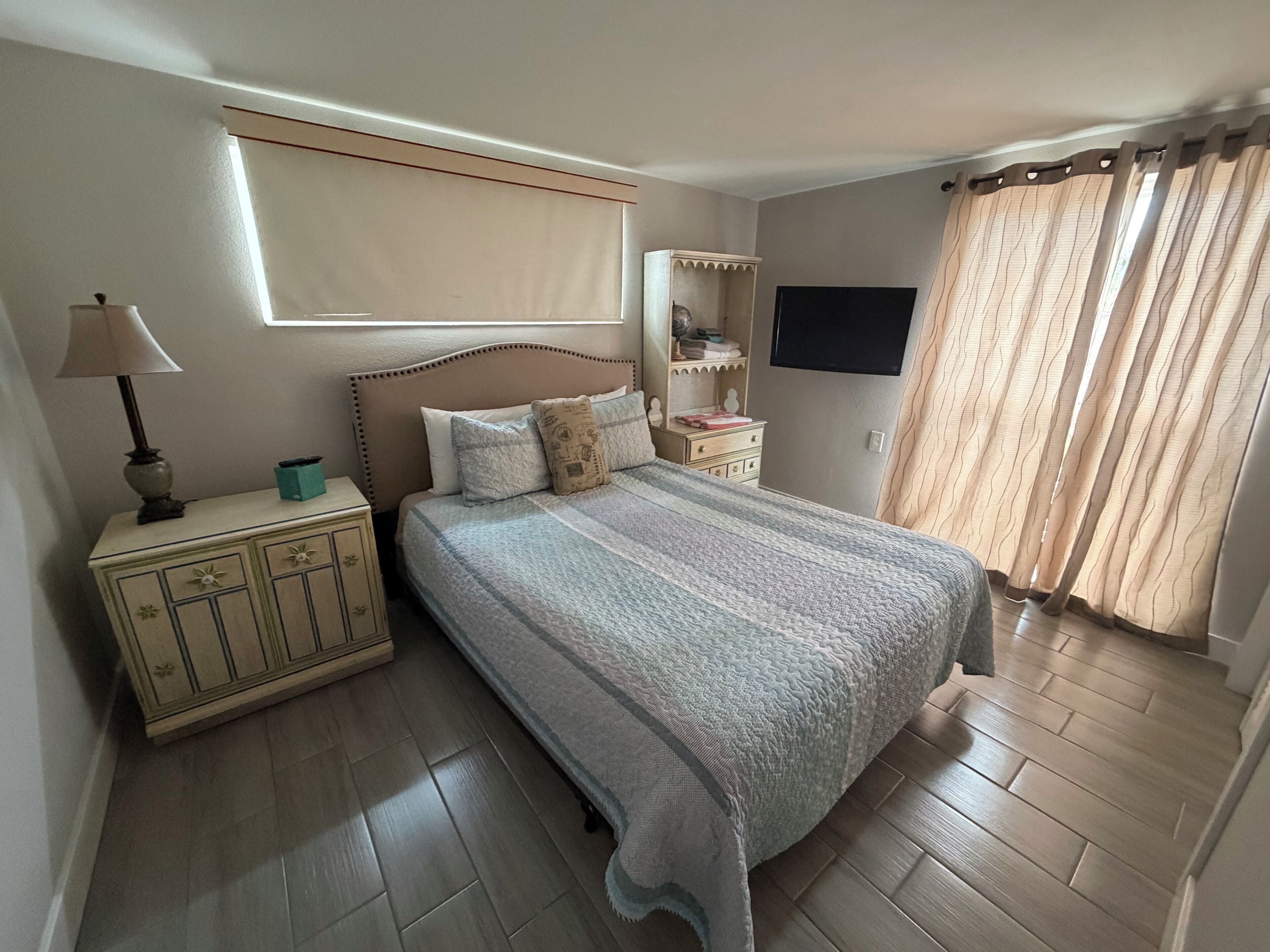 A bedroom with a bed, a bedside table with a lamp, a dresser, a TV, and a window with curtains.