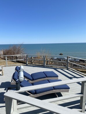Terraza de madera con dos sillones de descanso y una mesa, con vista a una costa rocosa y el mar.