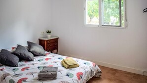 Een slaapkamer met een bed, kussens, een nachtkastje, een raam en uitzicht op bomen.