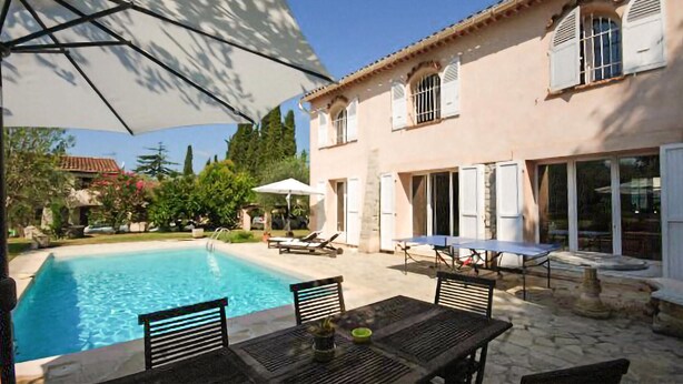 A poolside area with a table and chairs, a covered seating area, and a building with windows and shutters. - Lovely home in Mouans-Sartoux with swimming pool (Mouans-Sartoux)