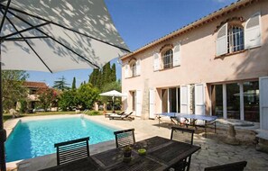 Un espace aménagé au bord de la piscine avec une table et des chaises, un coin salon couvert, et un bâtiment doté de fenêtres et de volets.