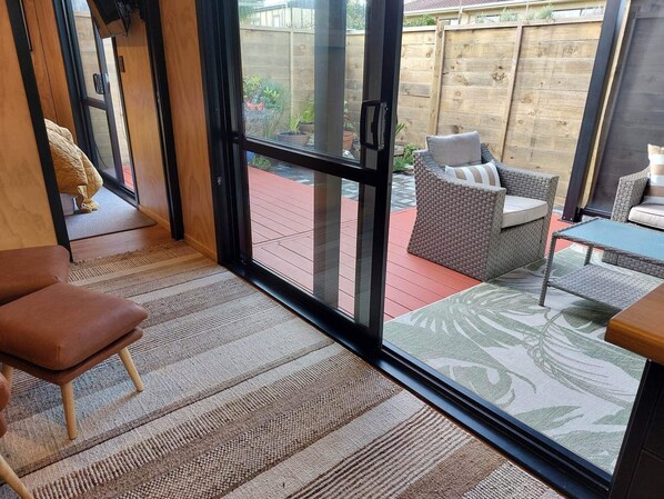 Ein Zimmer mit einer großen Schiebeglastür, die zu einer Terrasse führt. Innen befindet sich ein brauner Ledersessel, ein Korbsessel mit Kissen und ein kleiner Tisch mit Glasplatte.
