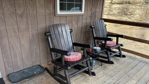 Two Adirondack chairs with red cushions on a wooden deck, a small table with a drink, and a window above.