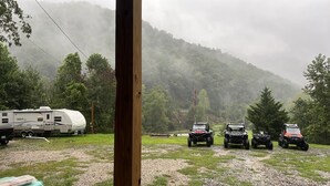 A campsite with a trailer and four ATVs parked on a gravel area, surrounded by trees and a forested hill.