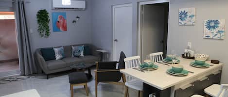 A modern kitchen with a dining area, grey walls, and a grey sofa with teal cushions.
