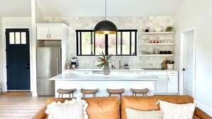A modern kitchen with white cabinetry, a central island, and bar stools. There is a brown leather sofa with decorative pillows in the foreground. - Lux Modern Barnhouse Cabin 1 minute from Hunter Mountain (Hunter)