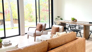 A modern living room with a large sofa, a dining area with a wooden table and chairs, and large windows offering a view of trees and a building outside. - Lux Modern Barnhouse Cabin 1 minute from Hunter Mountain (Hunter)