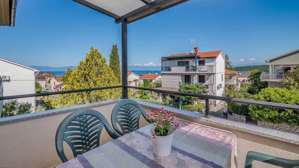 Een balkon met een tafel en stoelen, een bloempot en uitzicht op gebouwen en de zee.