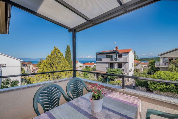A balcony with a table and chairs, a potted plant, and a view of buildings and the sea.