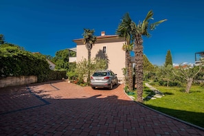 A house with a paved driveway, a parked car, and a well-maintained garden with palm trees.